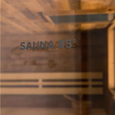 A sauna with a warm wooden interior. The glass door says "SAUNA 85°".