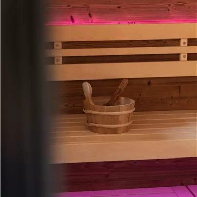 A cozy sauna with wooden shelves and a wooden bucket. Gentle, pink light creates a relaxing atmosphere.