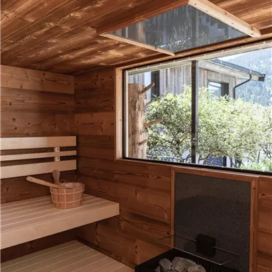 A cozy sauna room with wood paneling and a large window front. Inside, there is a bench and a heating stove.