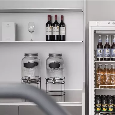 A modern bar with wine bottles, glasses, and two large storage jars. In the fridge, various non-alcoholic drinks are visible.