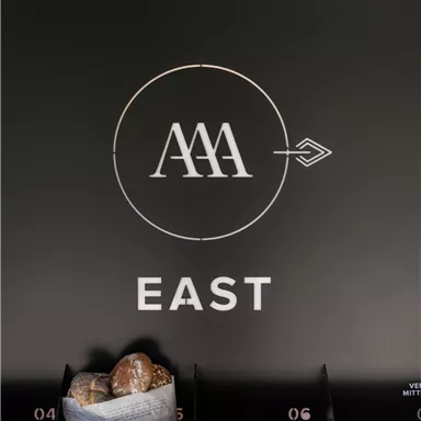 A black sign with the logo "AAA" and the word "EAST". Below are compartments with a basket containing some rolls.
