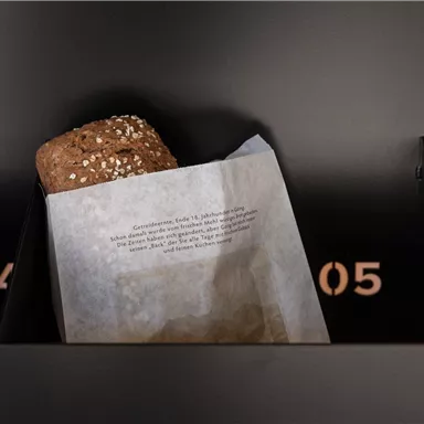 A roll with sesame in a paper bag lies in a dark gray compartment. In the background, two numbers are visible: 04 and 05.