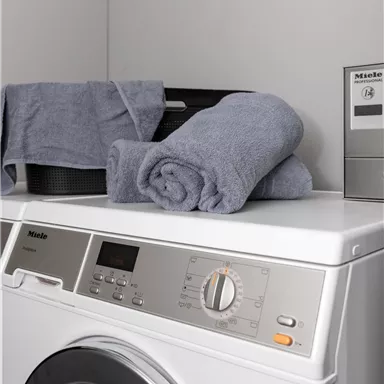 A modern washing machine with gray towels on top. In the background, there is a dark container and a control panel.