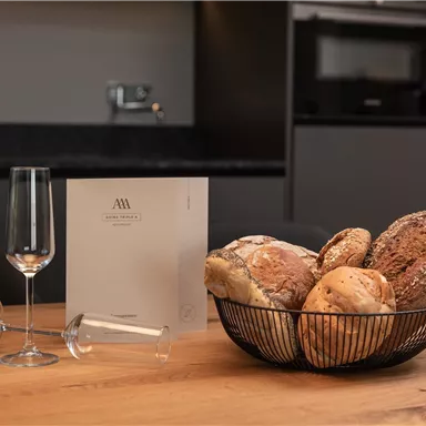 A basket of fresh bread is on a wooden table. Next to it are two glasses and a menu in a modern kitchen environment.
