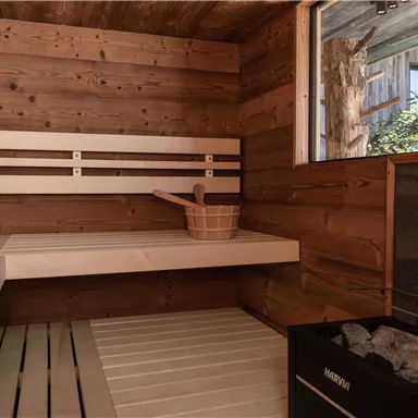 A cozy sauna made of light wood with benches and a sauna stone stove. Sunlight shines through the window and offers a view of the outdoors.