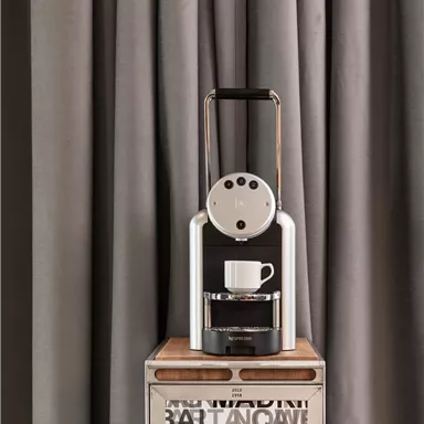 A modern coffee machine is on a table with a wooden surface. The background consists of gray curtains.