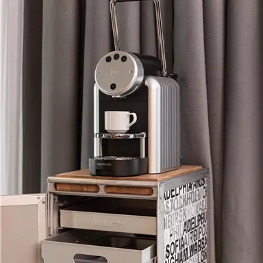 A modern coffee machine is sitting on a wooden table with a coffee cup. In the background, gray curtains are visible.