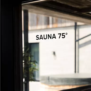 A glass window with the inscription "SAUNA 75°". In the background, a modern, bright building can be seen.