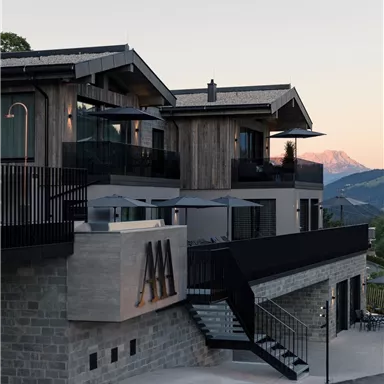 A modern building with wooden and stone elements, surrounded by mountainous landscape. The facade is stylish and inviting, ideal for a relaxing stay.