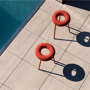 Two red chairs with an unusual design are placed on a light floor next to a pool. The shadows of the chairs are clearly visible on the floor.