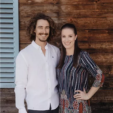 A couple stands in front of a wooden wall. Both are smiling friendly and are fashionably dressed.