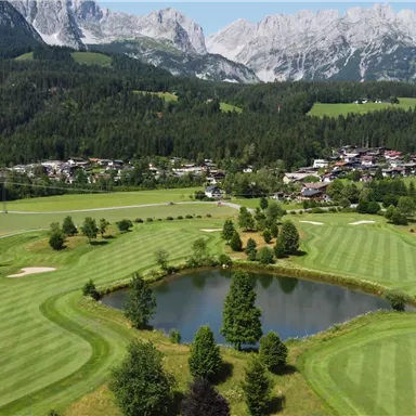 A picturesque golf course with well-maintained lawns and a small pond. In the background, majestic mountains and a charming village stretch out.