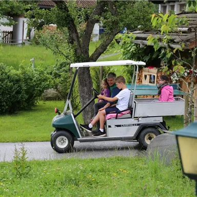 Ein kleiner Golfwagen fährt auf einem Weg durch eine grüne Landschaft. Darin sitzen mehrere Kinder, die Spaß haben.