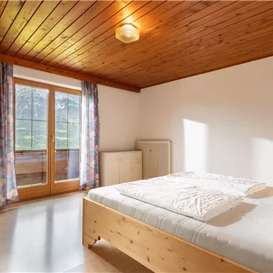 A bright bedroom with a wooden ceiling and large windows. There is a double bed and floral curtains.