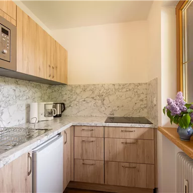 A modern kitchen with wooden furniture and a marbled countertop. The window offers a view of the surroundings and there is a vase of lilacs on the table.