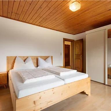 A cozy bedroom with a wooden double bed and two pillows. The room features warm wooden decor and a large mirror.