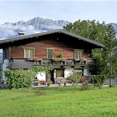A traditional wooden house surrounded by green areas and mountains. The atmosphere is peaceful and rural.