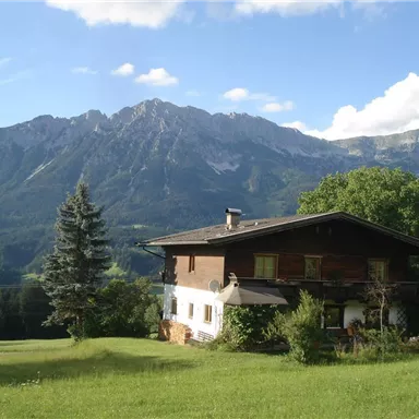 A beautiful house surrounded by green meadows and tall mountains. The sky is clear and sunny.
