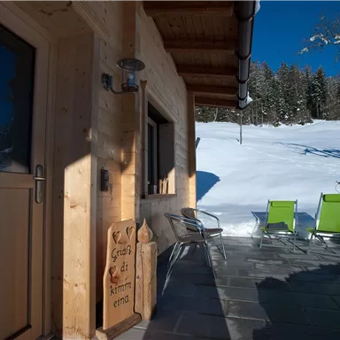 Eine gemütliche Holzveranda mit zwei grünen Stühlen in einer schneebedeckten Landschaft. Der Himmel ist klar und blau, umgeben von Bäumen.