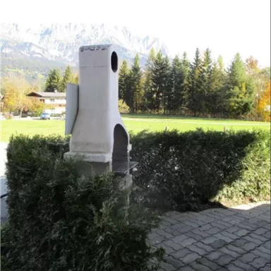 Ein Kamin aus Beton steht in einem Garten, umgeben von einem grünen Strauch. Im Hintergrund sind Bäume und Berge sichtbar.