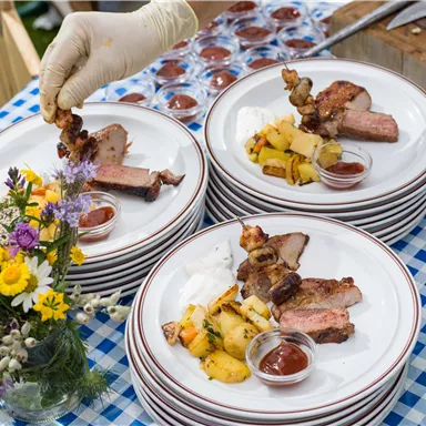 A table with several plates containing grilled meat and side dishes like potatoes and sauce. There are flowers in a vase as well.