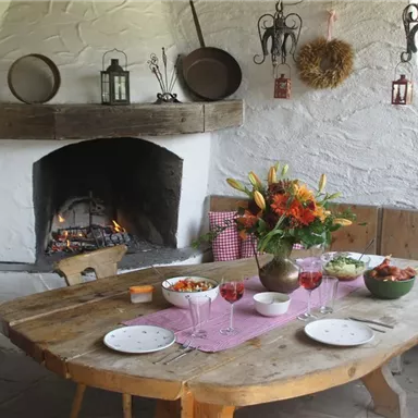 Ein rustikaler Essbereich mit einem großen Holztisch und einer Feuerstelle. Der Tisch ist gedeckt mit verschiedenen Gerichten und frischen Blumen in einer Vase.