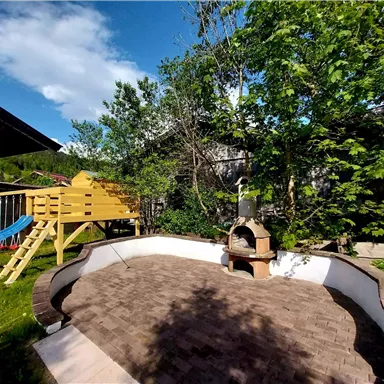Ein einladender Garten mit einer Steinterrasse und einem Grillplatz. Im Hintergrund steht ein Spielturm und es gibt viel grüne Vegetation.