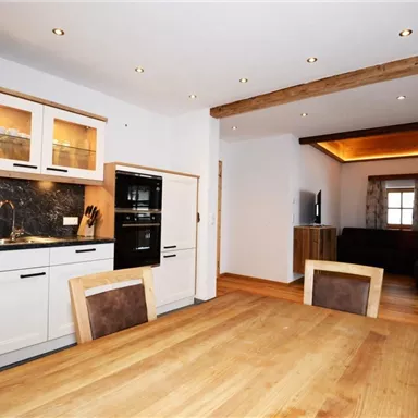 A modern kitchen with a large wooden dining table and high-quality appliances. In the background, there is a cozy living area with seating.