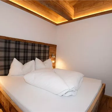 A cozy bedroom with a double bed and wooden details. The walls are kept simple, and a window with a green curtain lets in daylight.