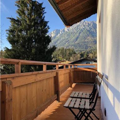 Ein Balkon mit Holzgeländer und zwei Stühlen. Im Hintergrund sind Berge und ein klarer blauer Himmel sichtbar.