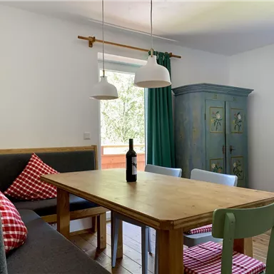 Ein gemütlicher Essbereich mit einem Holztisch und bunten Stühlen. Vor dem Fenster stehen grüne Vorhänge und eine Flasche Wein auf dem Tisch.