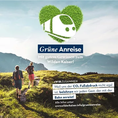Zwei Wanderer gehen auf einem Bergweg, umgeben von grüner Landschaft. Im Hintergrund sind Berge und ein blauer Himmel zu sehen, während das Motto "Grüne Anreise" hervorgehoben wird.