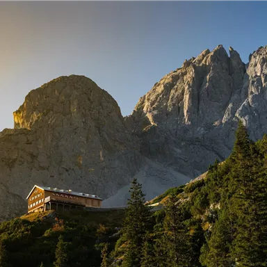 Ein malerisches Bergpanorama mit hohen Felsen und einer Hütte im Vordergrund. Die Sonne scheint sanft auf die Landschaft und die Bäume.