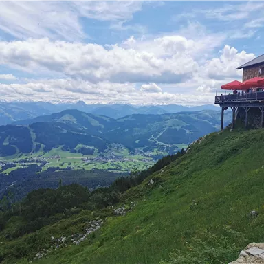 Eine Berglandschaft mit einem Restaurant, das über eine Terrasse mit roten Schirmen verfügt. Die grünen Wiesen und das weitläufige Tal bieten einen schönen Blick auf die umliegenden Berge.