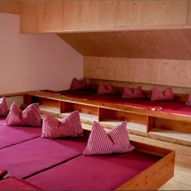 Ein gemütlicher Raum mit niedrigen Holzplattformen und roten Kissen. Die Wände sind mit Holz verkleidet und es gibt große Fenster, die Licht hereinlassen.