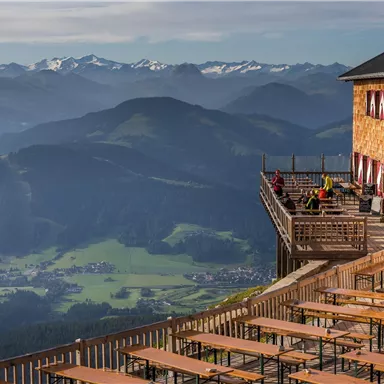 Eine Berghütte mit einer großen Terrasse und Holzstühlen bietet einen atemberaubenden Blick auf die umliegenden Berge. Die Landschaft ist grün mit verschiedenen Höhen und schneebedeckten Gipfeln im Hintergrund.