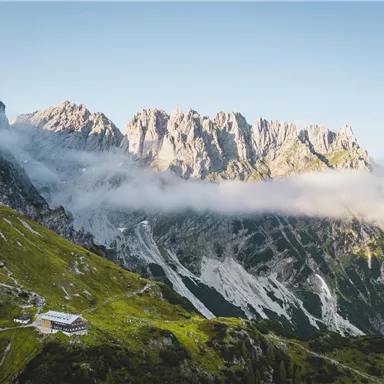 Eine beeindruckende Berglandschaft mit majestätischen Felsen und sanften Hügeln. Im Vordergrund ist eine kleine Hütte sichtbar, umgeben von üppigem Grün und Nebel.