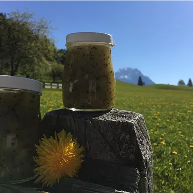 Two glasses with a green mixture are placed on a wooden board. In the background, a blooming meadow stretches under a clear sky and mountains.