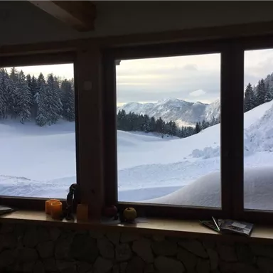 A winter view through a large window of snow-covered mountains and forests. The landscape is calm and picturesque with a gentle blanket of snow.