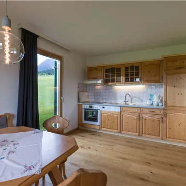 Eine helle Küche mit Holzverbauten und einem Tisch aus Holz. Durch das Fenster ist eine grüne Landschaft sichtbar.