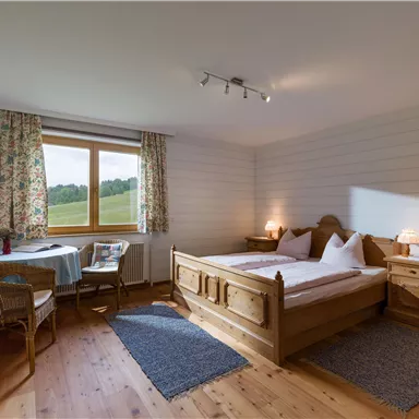 Ein gemütliches Schlafzimmer mit einem Doppelbett und zwei Nachttischen. Es gibt einen Tisch mit Stühlen und große Fenster, die viel Tageslicht hereinlassen.