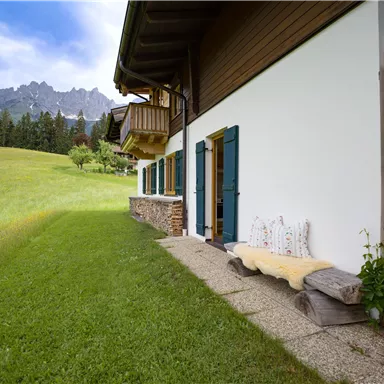 Ein idyllisches Haus in der Natur, umgeben von grünen Wiesen und Bergen im Hintergrund. Die Terrasse ist einladend mit Holzscheiten und einer bequemen Bank.
