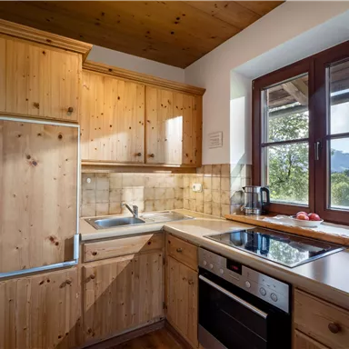 Eine moderne Küche mit Holzschränken und einem hellen Farbschema. Sie ist mit einem Herd, einer Spüle und einem Kühlschrank ausgestattet, die Licht durch die Fenster hereinlassen.