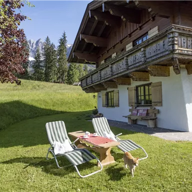 Ein gemütliches Chalet in einer grünen Landschaft, umgeben von Bäumen und Bergen. Vor dem Haus stehen zwei Liegen und ein Tisch mit Frühstück.
