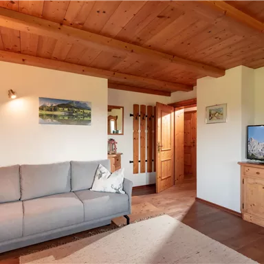 Ein gemütliches Wohnzimmer mit einem grauen Sofa und einer Holzdecke. Ein Fernseher und ein Regal aus Holz fügen sich harmonisch in die Einrichtung ein.