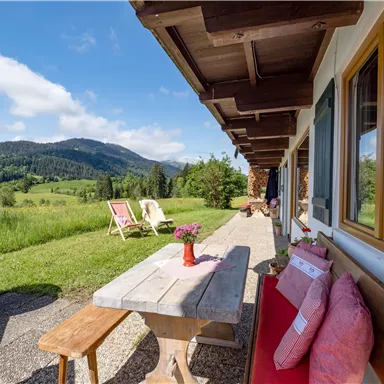Eine gemütliche Terrasse mit einem Holztisch und Sitzgelegenheiten. Im Hintergrund erstrecken sich grüne Wiesen und sanfte Hügel unter einem blauen Himmel.