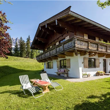 Ein gemütliches Alpenhaus mit einem Balkon und einem gepflegten Garten. Im Vordergrund stehen Liegestühle auf der grünen Wiese.