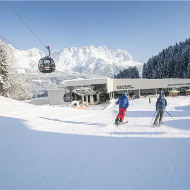 Two skiers are skiing on a snow-covered slope. In the background, the mountains and a cable car are visible.