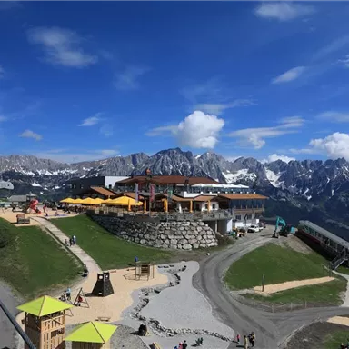 An impressive mountain landscape with snow-covered peaks and a cozy restaurant in the foreground. Guests and play areas can be seen on the premises.