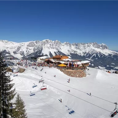 Ein schöner winterlicher Skiort mit schneebedeckten Bergen und einem klaren blauen Himmel. Skifahrer nutzen die Pisten, während eine hochgelegene Hütte zum Verweilen einlädt.